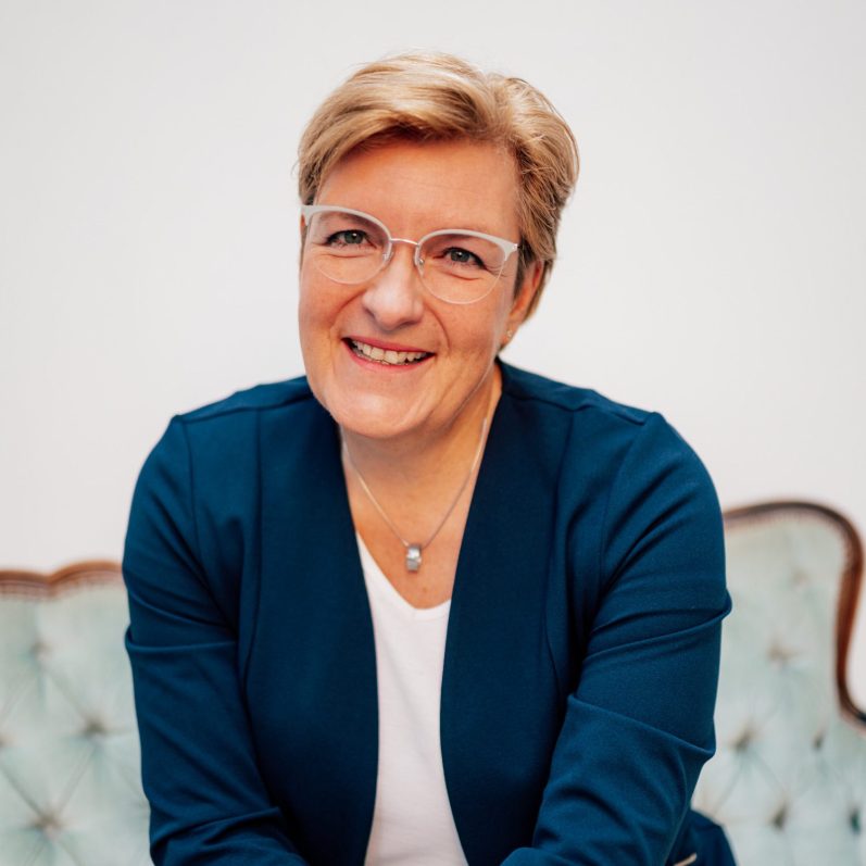 Lächelnde Frau mit Brille, trägt einen blauen Blazer, sitzt auf einem Sofa.