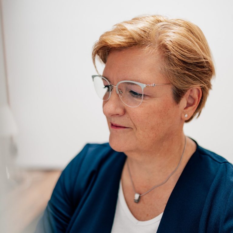 Frau mit blonden Haaren und Brille, nachdenklich und in einem blauen Blazer.
